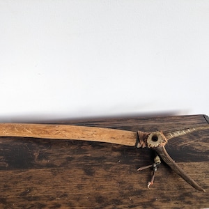 Puede incluir: Un tomahawk de madera hecho a mano con un mango largo y plano y una hoja puntiaguda. La hoja está unida al mango con un envoltorio marrón. También hay pequeñas plumas y detalles de cuero. El tomahawk descansa sobre una superficie de madera oscura.