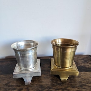 May include: Two metal stocking holders. One is silver, the other is gold. Each has a bucket-shaped top and a square base with a hook for hanging. They are displayed on a dark wooden surface.