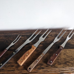 Op de afbeelding: Een verzameling van zes vintage vleesvorken met roestvrijstalen tanden en houten handvatten. De handvatten variëren in kleur van donkerbruin tot licht houtnerf. De vorken zijn gerangschikt op een donker houten oppervlak.