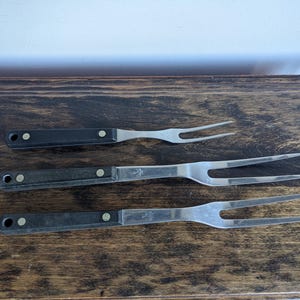 May include: Three silver grilling forks with black plastic handles. The forks are arranged in a row on a wooden surface.