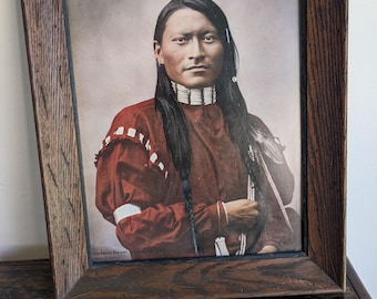 May include: A framed portrait of a Native American man with long black hair, wearing a red and white beaded shirt and a beaded necklace. The portrait is in a dark wood frame.