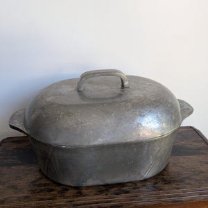 May include: A vintage, oval-shaped, cast-iron Dutch oven with a lid. The pot is a dull silver color with a handle on the lid and two side handles. The oven sits on a dark wooden table against a white wall.