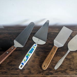 May include: Four vintage serving utensils on a dark wood surface. Includes two cake servers with stainless steel blades and wooden handles, a spatula with a wooden handle, and a small serving spoon.