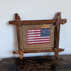 Pode incluir: Uma bandeira americana emoldurada. A bandeira tem listras vermelhas e brancas e um campo azul com estrelas brancas. A moldura é de madeira marrom escura com detalhes de folhas esculpidas em cada canto.