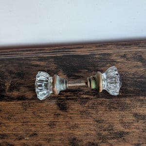 May include: A door knob set featuring two clear, fluted glass knobs connected by a central metal spindle. Each glass knob is attached to a circular, weathered brass-toned metal base with visible patina and a small set screw on the neck.