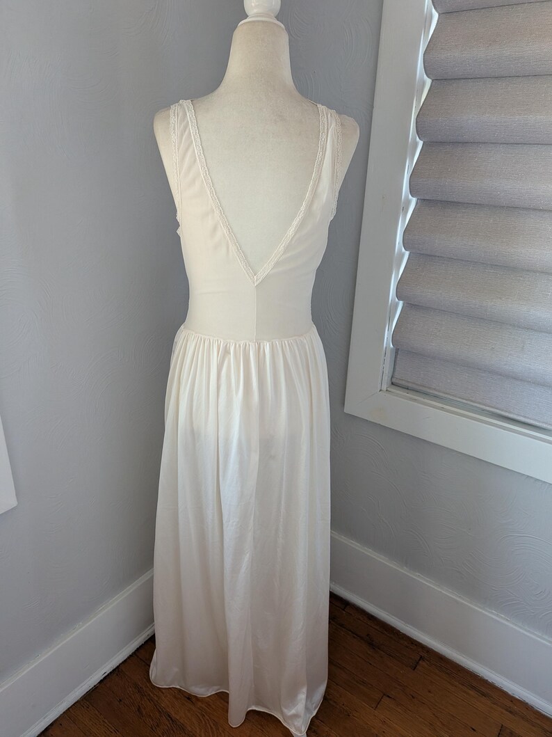 May include: Cream-colored vintage nightgown on a mannequin. The gown features a V-neckline with lace trim and a flowing, floor-length skirt. The fabric appears to be a soft, silky material. The back of the gown is visible, showcasing the delicate design.