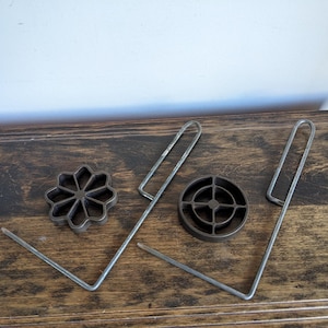 May include: A collection of vintage metal cookie cutters and a pair of metal handles, arranged on a wooden surface. The cutters include a flower shape and a circular design. The handles are bent into a V-shape.