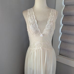 May include: Ivory-colored vintage nightgown with a V-neckline and delicate lace trim. The sleeveless gown features lace detailing on the bodice and a flowing skirt. A small bow accents the neckline. The nightgown is displayed on a mannequin.