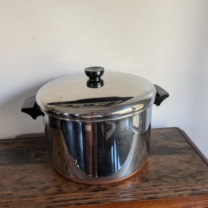 Puede incluir: Una olla de acero inoxidable pulido con tapa a juego, que cuenta con dos asas laterales negras y un pomo negro en la tapa. La olla tiene un cuerpo cilíndrico con una superficie reflectante y una base de color cobre, con acabado metálico.