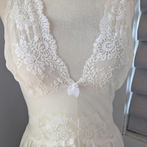 May include: Ivory-colored lingerie with a V-neckline and delicate lace detailing. The sleeveless garment features floral lace accents on the bodice and a flowing skirt. A small bow adorns the center of the bust.
