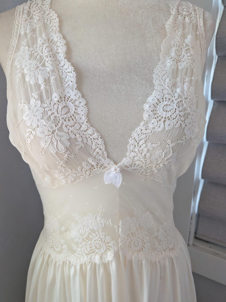 May include: Ivory-colored lingerie with a V-neckline and delicate lace detailing. The sleeveless garment features floral lace accents on the bodice and a flowing skirt. A small bow adorns the center of the bust.
