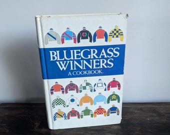 Könnte beinhalten: Ein weißes Kochbuch mit dem Titel "Bluegrass Winners: A Cookbook" mit einem blauen Rand und einem Design aus bunten Jockey-Seiden auf dem Cover.