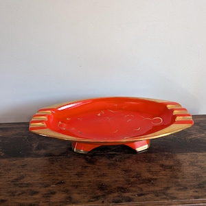 Può includere: Un posacenere vintage rosso acceso con finiture dorate. Di forma ovale, poggia su tre piedini. Il posacenere presenta incavi per sigarette, un design retrò.