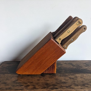 Può includere: Un blocco di coltelli in legno con una finitura marrone rossiccia contiene un set di cinque coltelli con manici in legno. I coltelli hanno dettagli in ottone e il manico di un coltello ha la scritta "CHIEF" stampata.