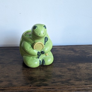 May include: A green ceramic turtle figurine holding a yellow tennis racket. The turtle is sitting on a wooden surface.