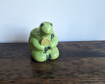 Puede incluir: Una figurita de tortuga de cerámica verde que sostiene una raqueta de tenis amarilla. La tortuga está sentada sobre una superficie de madera.