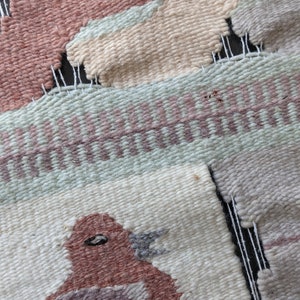 Pode incluir: Uma tape&ccedil;aria tecida para pendurar na parede com um p&aacute;ssaro, dois animais e um padr&atilde;o geom&eacute;trico. O p&aacute;ssaro &eacute; marrom com um bico e um olho cinza. Os animais s&atilde;o bege e marrons. O padr&atilde;o geom&eacute;trico &eacute; verde, rosa e branco.