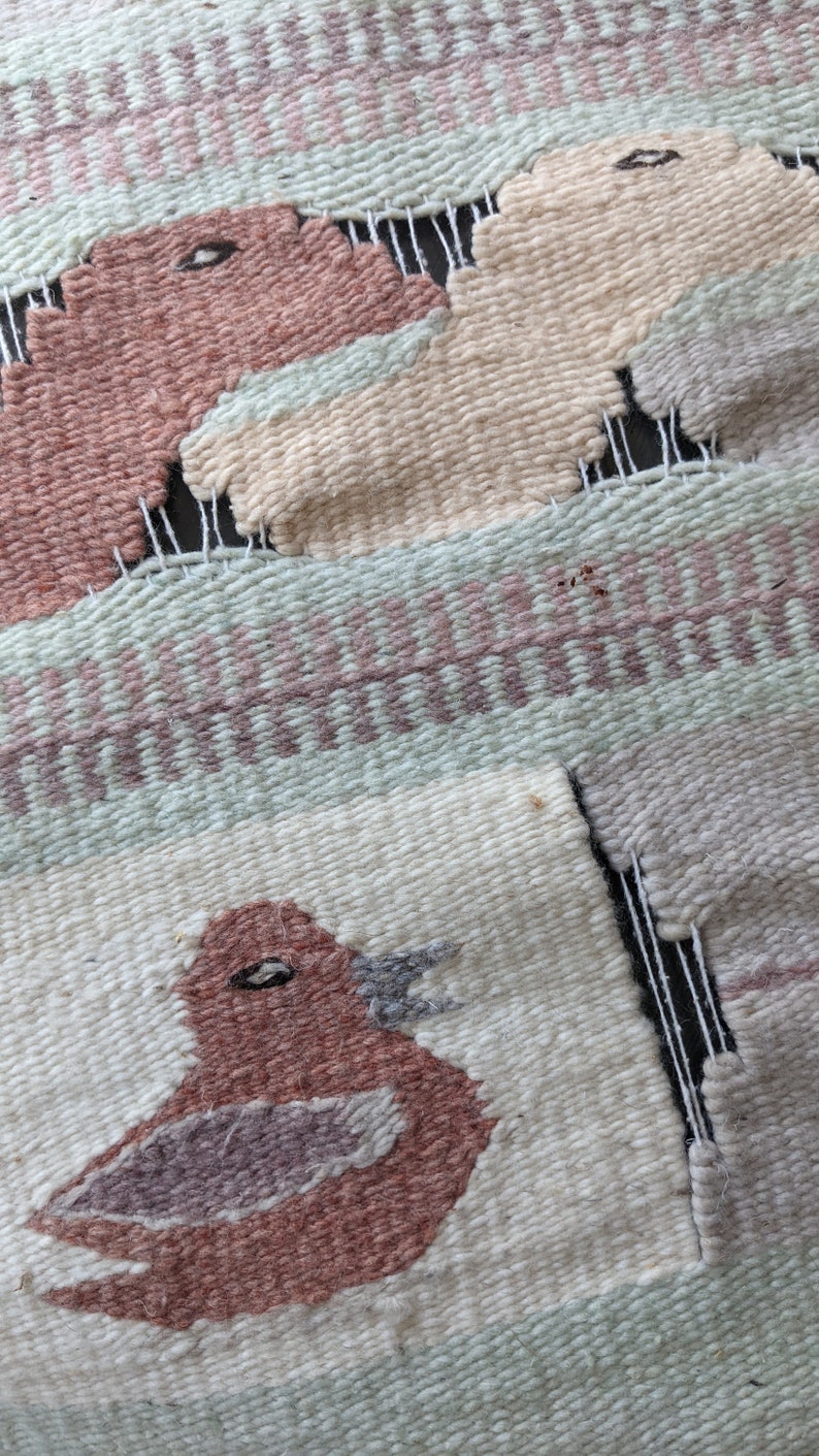 Pode incluir: Uma tape&ccedil;aria tecida para pendurar na parede com um p&aacute;ssaro, dois animais e um padr&atilde;o geom&eacute;trico. O p&aacute;ssaro &eacute; marrom com um bico e um olho cinza. Os animais s&atilde;o bege e marrons. O padr&atilde;o geom&eacute;trico &eacute; verde, rosa e branco.