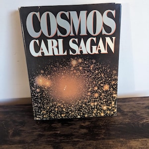 May include: A hardcover book titled "COSMOS" by Carl Sagan. The book cover is black with silver lettering and a cosmic design featuring a central bright orange burst surrounded by numerous smaller white and gold dots.