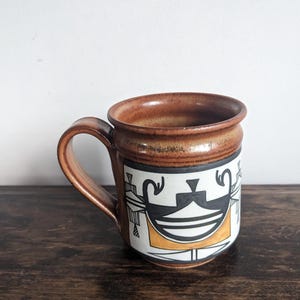 以下が含まれることがあります： 取っ手付きの茶色のセラミックマグカップ。マグカップは、黒、白、オレンジの幾何学模様が特徴です。マグカップは、濃い木製の表面に置かれています。