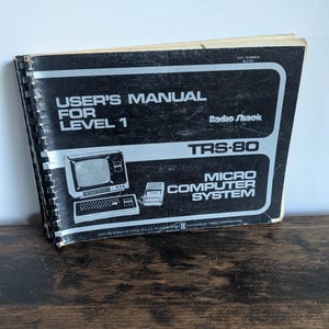 May include: A black and white spiral-bound user's manual for the Radio Shack TRS-80 micro computer system. The cover features the text "USER'S MANUAL FOR LEVEL 1", "Radio Shack", "TRS-80", and "MICRO COMPUTER SYSTEM" with a graphic illustration.