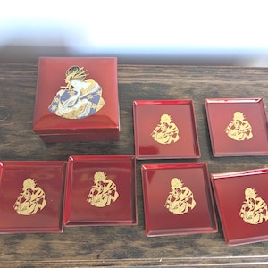 May include: A red lacquered box and six square coasters. Each piece features a gold illustration of a figure in traditional attire. The box and coasters are arranged on a dark wooden surface.