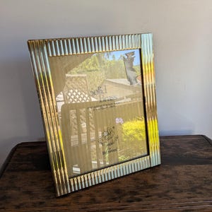 May include: A rectangular picture frame made of solid brass with a vertical ribbed texture along the border. The glass insert features printed text stating Solid Brass, hand polished & lacquer finished, 4 inch by 6 inch / 10cm by 15cm, and Bewon.