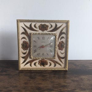 Op de afbeelding: Een vintage gouden klok met een witte wijzerplaat en zwarte Romeinse cijfers. De klok heeft een bloemmotief met gouden accenten en een bruine achtergrond.