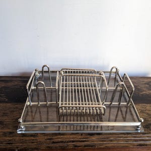 Puede incluir: Un organizador de escritorio plateado con múltiples compartimentos. El organizador tiene una base rectangular con una superficie de vidrio transparente y presenta varias secciones verticales y una sección central con un patrón de rejilla.