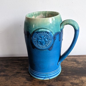 May include: A blue and green ceramic beer stein with a handle. The stein has a circular emblem that reads "The Renaissance Faire, Pleasure + Fame".