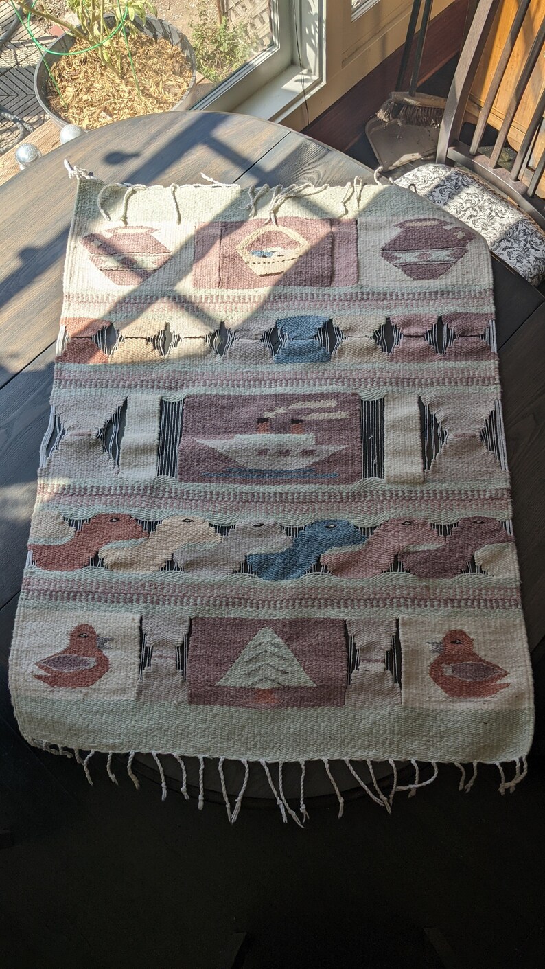 Pode incluir: Uma tape&ccedil;aria de parede tecida com um design geom&eacute;trico que apresenta p&aacute;ssaros, barcos e formas abstratas em tons de marrom, azul e branco. A tape&ccedil;aria tem uma borda de franjas.