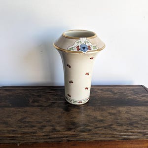 May include: A white ceramic vase with a gold rim and floral design. The vase has a flared top and a narrow base. It is decorated with small red dots and a blue flower.
