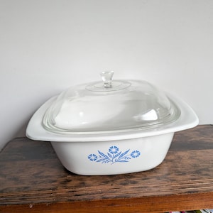 Puede incluir: Fuente de horno blanca con tapa de cristal transparente. La fuente tiene forma cuadrada con bordes redondeados y un estampado floral azul. La tapa tiene un pequeño tirador de pomo. La fuente está sobre una superficie de madera.