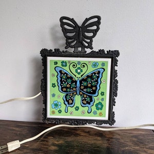 Puede incluir: Una lámpara de pared de mariposa de hierro fundido negro con una baldosa de mariposa verde y azul. La baldosa tiene un diseño floral y la lámpara tiene un cable blanco.