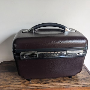 May include: A vintage brown travel case with a black handle and silver-toned hardware. The case has a textured exterior and sits on a wooden surface. The case is a classic piece of luggage.