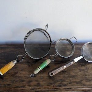 Może przedstawiać: Trzy vintage metalowe sitka z drewnianymi uchwytami. Największe sitko ma żółty i czarny uchwyt, średnie sitko ma zielony i biały uchwyt, a najmniejsze sitko ma brązowy uchwyt.