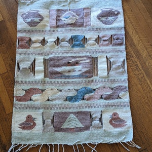 Pode incluir: Um tapete tecido com um padr&atilde;o geom&eacute;trico em tons de marrom, bege e azul. O tapete tem uma borda de franjas e apresenta p&aacute;ssaros estilizados e outros desenhos abstratos.