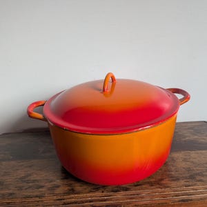 May include: A round enamelled cast iron Dutch oven featuring a vibrant gradient finish that transitions from a deep red at the base and lid rim to a bright orange in the centre. It includes two side handles and a matching lid with a loop handle.