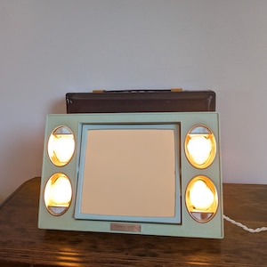 May include: A rectangular mint green vanity mirror with a central square reflective surface. Four oval-shaped light fixtures are arranged vertically on the left and right sides, each containing a glowing light bulb. A small label is centered below.