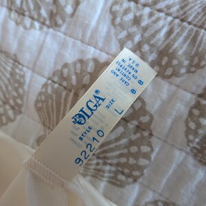May include: Cream-colored lingerie with a tag that reads "OLGA" and "SIZE L". The tag also includes the style number "92210" and "MADE IN U.S.A.". The lingerie is on a white and tan seashell patterned fabric.