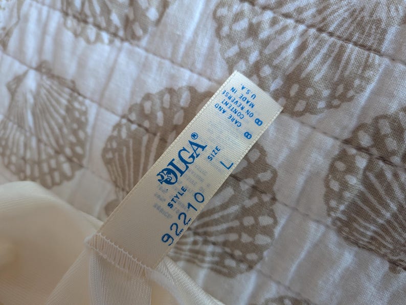 May include: Cream-colored lingerie with a tag that reads "OLGA" and "SIZE L". The tag also includes the style number "92210" and "MADE IN U.S.A.". The lingerie is on a white and tan seashell patterned fabric.