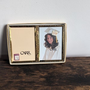 Peut inclure: Un cadre photo double avec une bordure dorée. Le cadre est en plastique et a un fond blanc. Le côté gauche du cadre est vide, tandis que le côté droit contient une photo d'une jeune femme portant une toque et une robe de graduation. La photo est en couleur et a un fond blanc. Le cadre porte l'inscription "CARR".