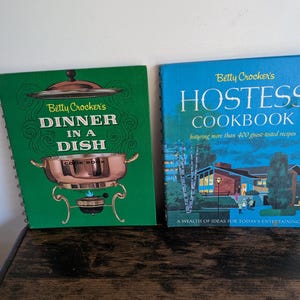 May include: Two vintage Betty Crocker cookbooks. The green book is titled "Dinner in a Dish" and features a copper chafing dish illustration. The blue book is titled "Hostess Cookbook" and depicts a mid-century house with people gathering outside.