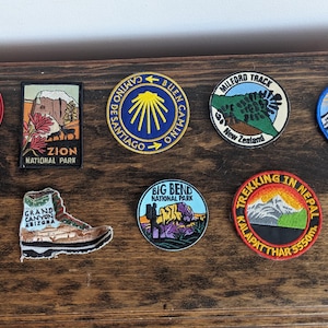 Può includere: Una collezione di toppe ricamate colorate che rappresentano vari parchi nazionali, montagne e monumenti. Le toppe sono esposte su una superficie di legno. Alcune delle toppe includono i nomi "Jackson Hole Wyoming", "Zion National Park", "Big Bend National Park", "Milford Track", "Mt. Whitney", "Trekking in Nepal", e "Mauna Kea Observatories".