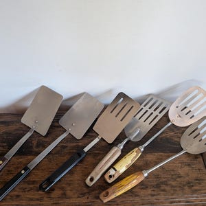 May include: A collection of six metal spatulas with varying handle designs. Three spatulas feature solid metal heads, while the other three have slotted heads. The handles include dark wood, black plastic, and weathered yellow painted wood finishes.