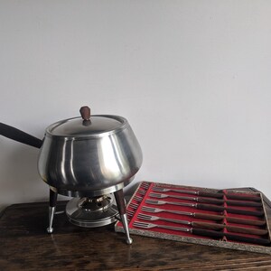 Pode incluir: Um conjunto de fondue vintage com uma panela de aço inoxidável, cabo e tampa de madeira. O conjunto inclui um suporte com pernas de madeira e uma caixa de garfos de fondue com cabos de madeira. A panela mede aproximadamente 20 cm de diâmetro.