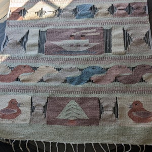 Pode incluir: Uma tape&ccedil;aria de parede tecida com um esquema de cores verde, marrom e rosa. A tape&ccedil;aria apresenta uma variedade de formas geom&eacute;tricas e desenhos, incluindo p&aacute;ssaros, &aacute;rvores e barcos. A tape&ccedil;aria tem uma borda de franjas.