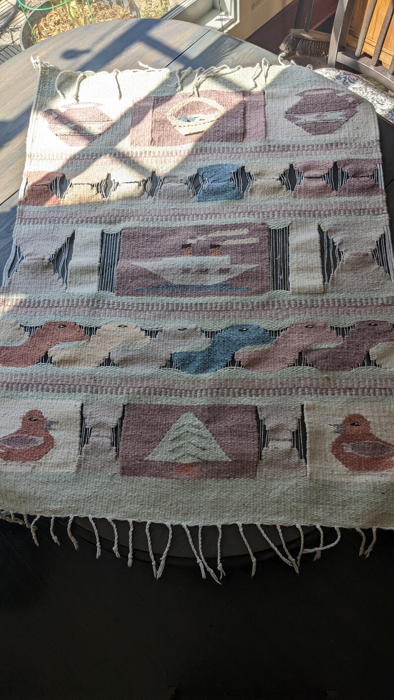 Pode incluir: Uma tape&ccedil;aria de parede tecida com um esquema de cores verde, marrom e rosa. A tape&ccedil;aria apresenta uma variedade de formas geom&eacute;tricas e desenhos, incluindo p&aacute;ssaros, &aacute;rvores e barcos. A tape&ccedil;aria tem uma borda de franjas.