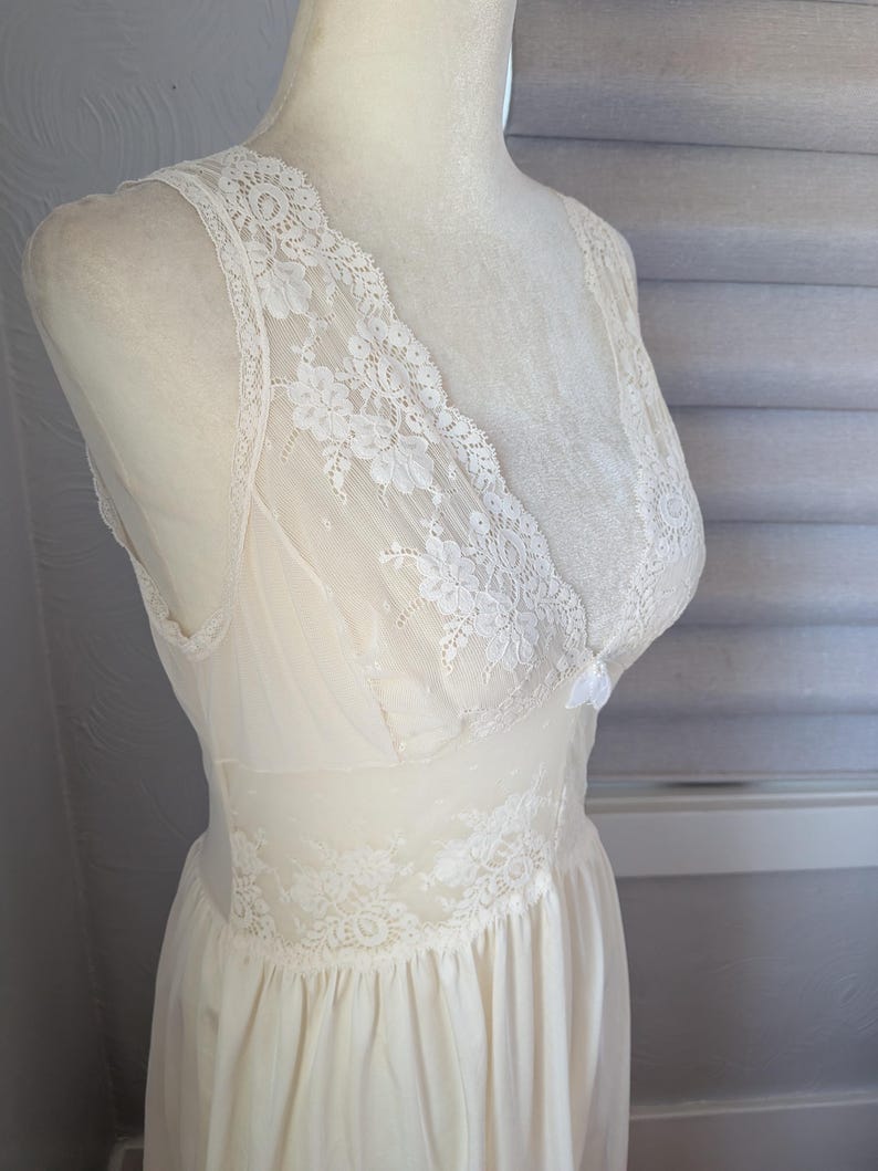May include: Ivory-colored vintage slip dress with delicate lace detailing. The dress features a V-neckline and lace trim along the bodice and straps. The skirt is flowing and gathered at the waist, creating a classic silhouette.