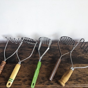 May include: A collection of vintage potato mashers with wooden and plastic handles. The mashers have different head designs, including wire grids and looped wires. The handles vary in color, with shades of brown, yellow, and green.